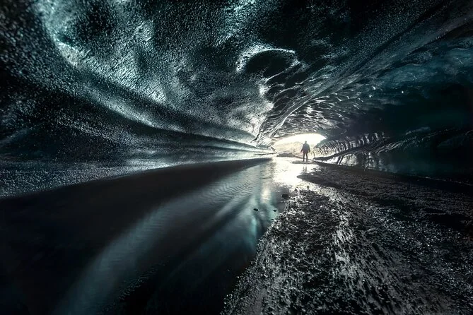 A dark ice tunnel with reflections on the floor and a small figure far away